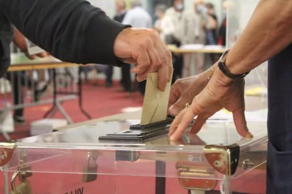 Une main met dans une urne transparente une enveloppe de vote